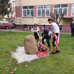 Troje uczniów z klasy ósmej składa kwiaty  i pali znicze  pod Dębem Pamięci upamiętniającym jedną z ofiar Zbrodni Katyńskiej aspiranta Policji Państwowej Bolesława Stasieckiego. W tle Dyrektor szkoły oraz  ks Proboszcz 