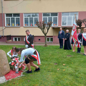 Troje uczniów z klasy ósmej składa kwiaty  i pali znicze  pod Dębem Pamięci upamiętniającym jedną z ofiar Zbrodni Katyńskiej aspiranta Policji Państwowej Bolesława Stasieckiego. W tle Dyrektor szkoły, ks Proboszcz i Poczet sztandarowy.